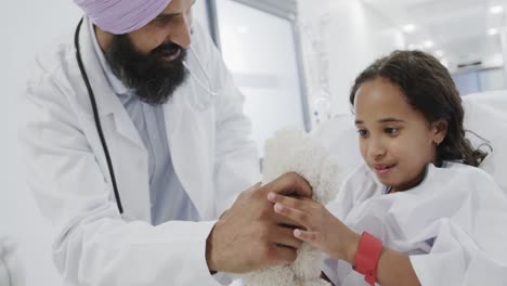 happy biracial doctor playing with sick girl patient with teddy bear in hospital bed in slow motion