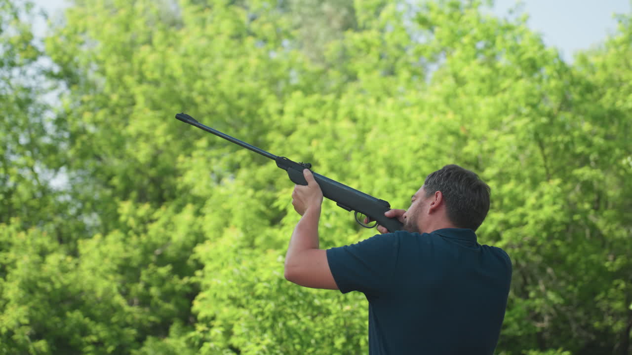 Man bending pneumatic rifle while aiming upward to chase bird from garden, summer backyard with green trees, cautious pest control protecting crops and wildlife balance in rural setting