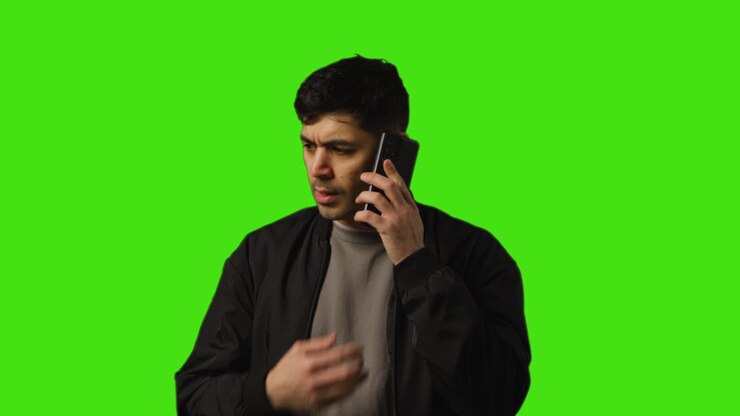Unhappy Young Man Answering Call On Mobile Phone Standing Against Green Screen Studio Background