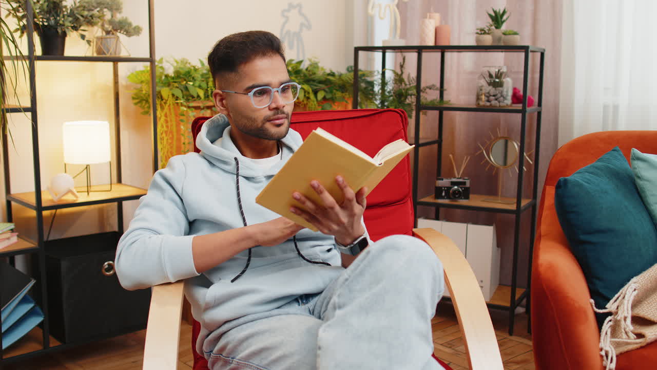 Indian man reading interesting book turning pages smiling enjoying literature taking a rest on chair