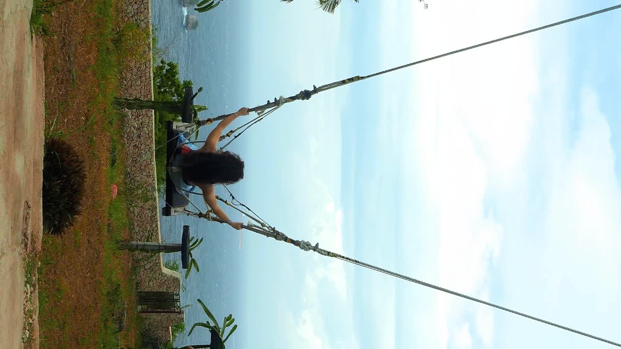 disparo vertical en cámara lenta de una mujer joven y atractiva vestida con falda azul cielo y camiseta roja en un columpio de bali en nusa penida en bali, indonesia con vista a las palmeras, los árboles y el mar