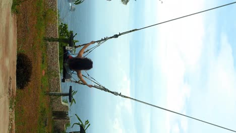 vertical slow motion dolly shot of a young attractive woman dressed in sky blue skirt and red top on a bali swing on nusa penida in bali indonesia with view of palm trees, trees and the sea