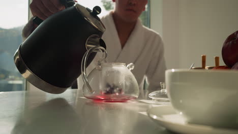 un caballero vierte agua hirviendo en una tetera. un hombre cariñoso llena la tetera con líquido calentado para empapar el té de frutas en la mesa de comedor en las vacaciones de verano