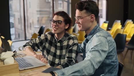 A couple of two young people wearing glasses, sitting next to each other, talking with someone through video chat. Enjoying time spent together.