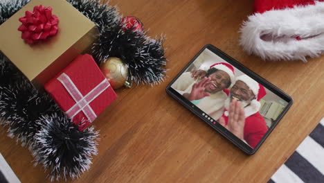 una pareja de adultos africanos-americanos sonrientes con sombreros de papá noel en una llamada de video de navidad en una tableta