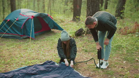 young man pumps air into tent bag using hand pump as woman smiles and holds fabric, surrounded by trees and forest tent in background during outdoor camping preparation in nature