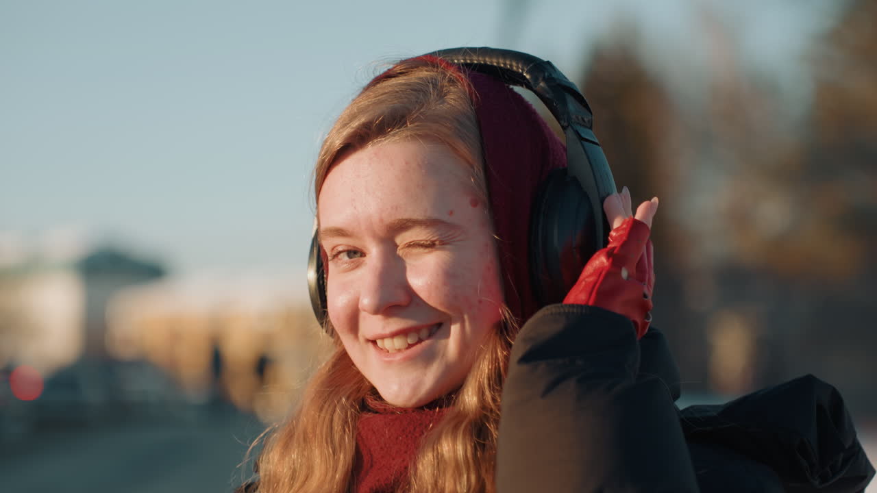Primo piano di giovane donna che sorride alla telecamera, cuffie sulle orecchie, guanti rossi e dolcevita caldo visibili, luce dell'ora d'oro, sfondo di parcheggio, espressione allegra e coinvolgente e gesto della mano.