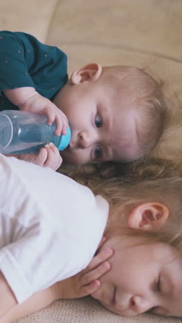 funny little baby looks at sleeping sister and drinks water from bottle on large bed in spacious room closeup