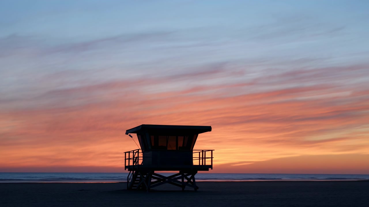 Et livreddertårn står i silhuet mod en levende solnedgang på en rolig strand ved skumring, der fanger et fredfyldt og roligt øjeblik med et vidvinkelbillede fra strandniveau. Metaltårnet