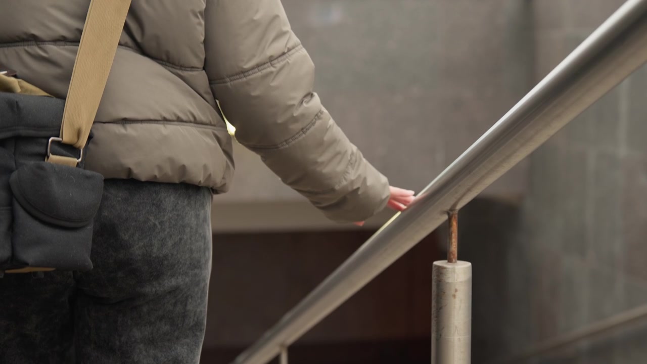 Kvinde går op ad trapper. Kvinde med taske bestiger trappe. Person i frakke bruger metalgelænder til opstigning. Kvindelig person navigerer marmortrappe med gelænder og taske i bymiljø.