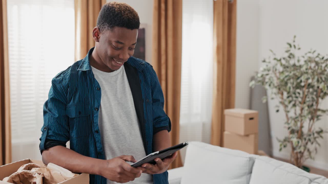 A young boy hangs out in his new apartment, unpacking cardboard boxes left over from the move, browsing the web on tablet, social media, laughing at memes, browsing inspiration