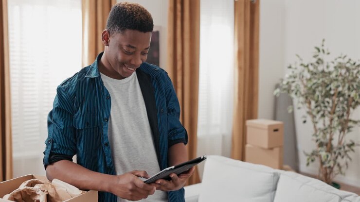 A young boy hangs out in his new apartment, unpacking cardboard boxes left over from the move, browsing the web on tablet, social media, laughing at memes, browsing inspiration