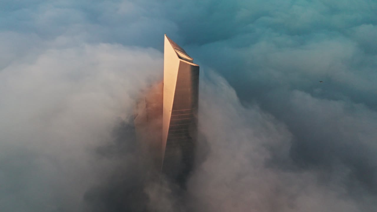 Cinematic drone shot of a modern skyscraper emerging above a sea of clouds and mist at sunrise, glowing in golden-hour light with dramatic shadows and soft haze