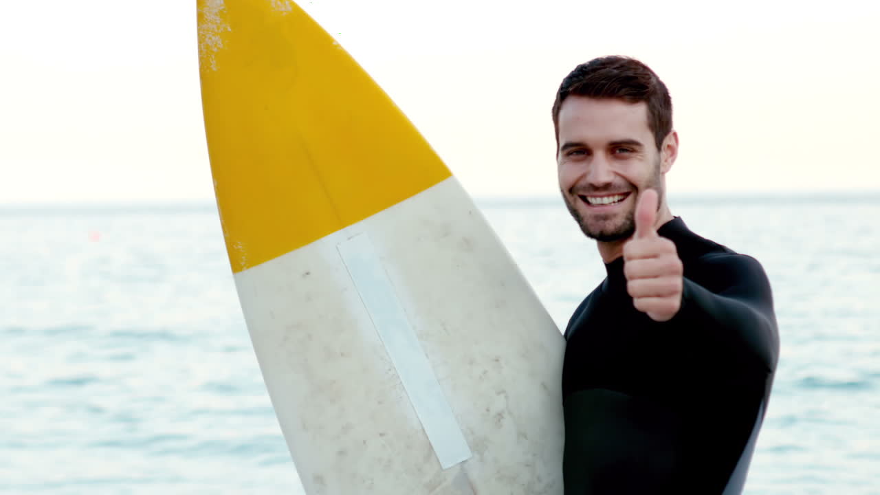 Uomo in muta con una tavola da surf che fa il gesto del pollice in su