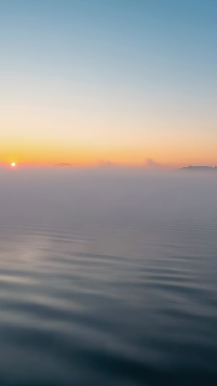 Vertical video: Rising sun bathing calm seascape at dawn, warming rippled water and low fog