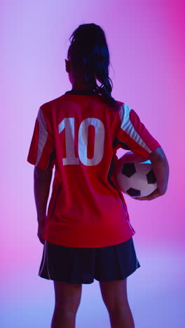 video vertical de vista trasera de una jugadora de fútbol o fútbol femenina con la camiseta del equipo sosteniendo la pelota bajo el brazo en un estudio disparado en clave baja contra una iluminación mixta colorida