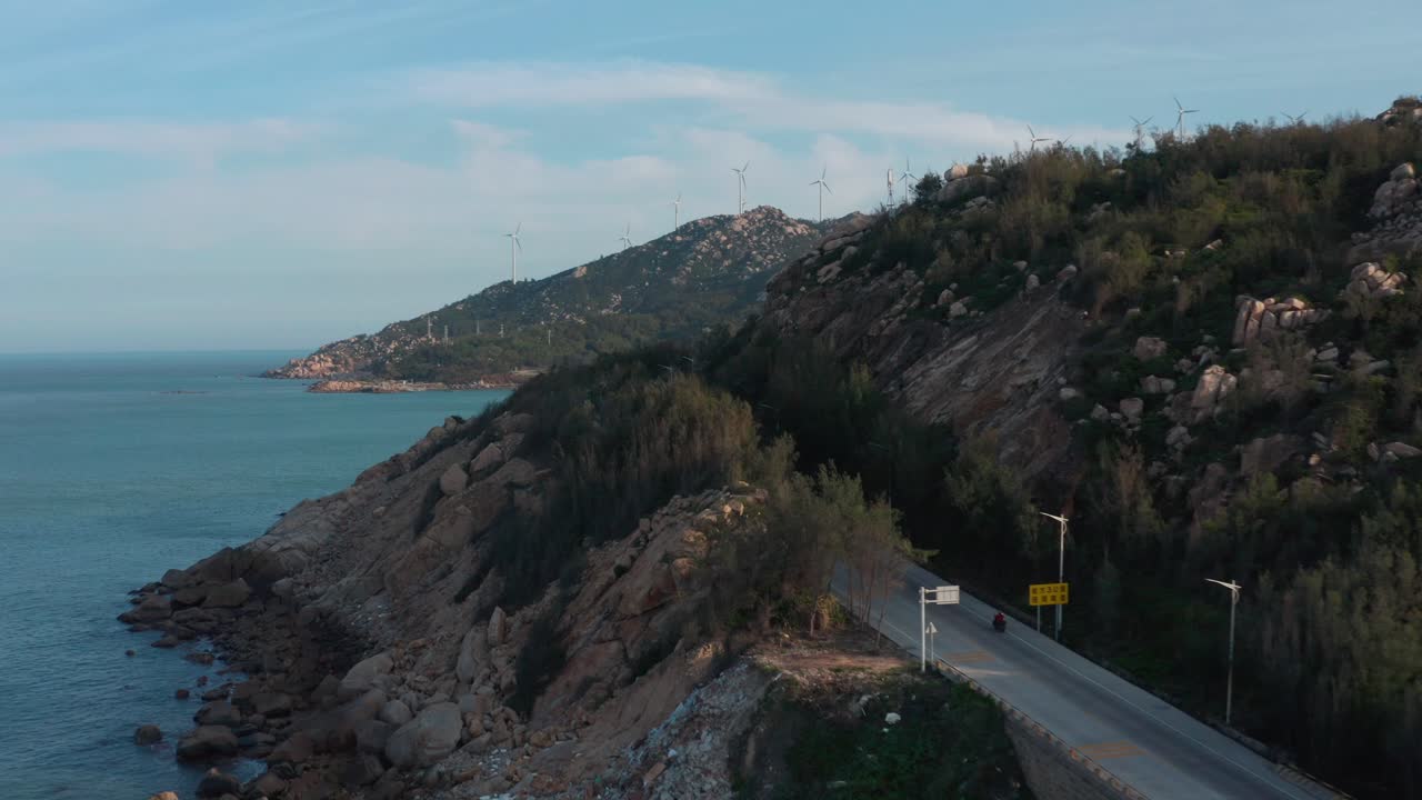 пролет над приморской дорогой с изображением ветряной электростанции на вершине горного хребта вдали.