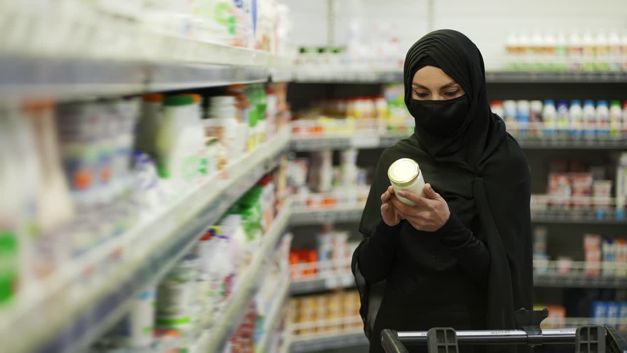 mujer con hijab y máscara protectora haciendo compras, toma el producto del estante en una sección de leche