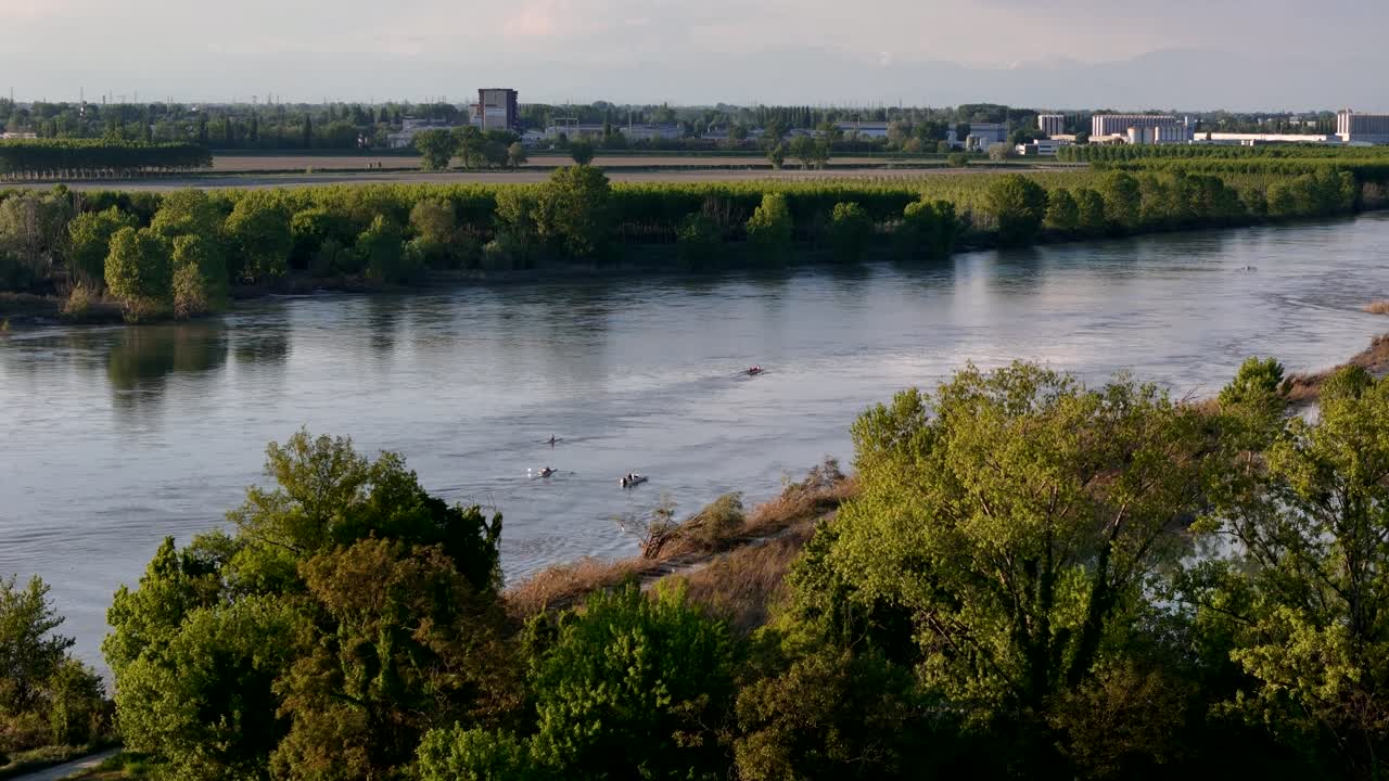 Ripresa aerea in slow-motion con allontanamento che rivela l'ampio fiume Po, delimitato da alberi verdi e lussureggianti, con una squadra di canottaggio che si allena sulle acque calme a Olza, Piacenza, nel nord Italia, con Cremona dall'altra parte.