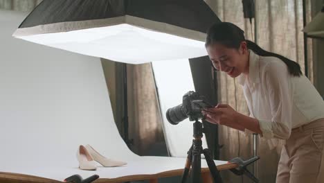 asian female photographer comparing photos on a smartphone to camera while taking photos of women's shoes in home studio