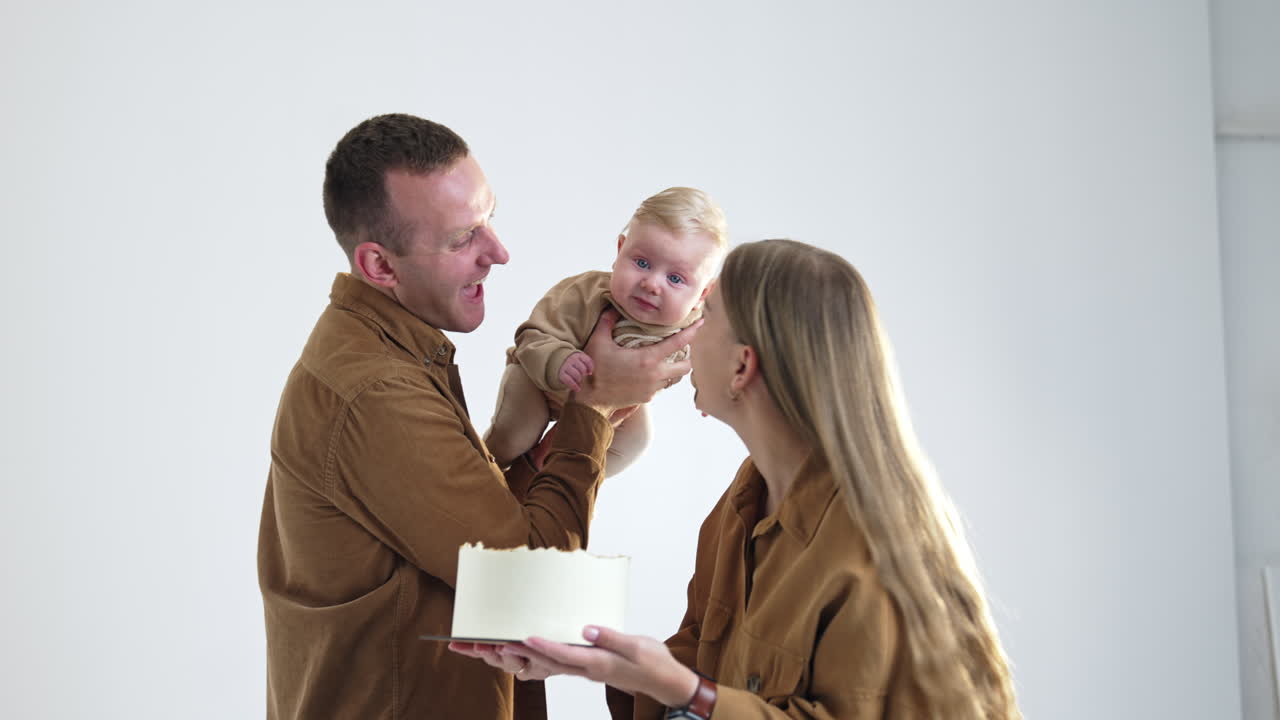 Happy parents wearing brown shirts celebrate their newborn's four months. Dad holds a cute infant son and mom holds a cake. White backdrop.