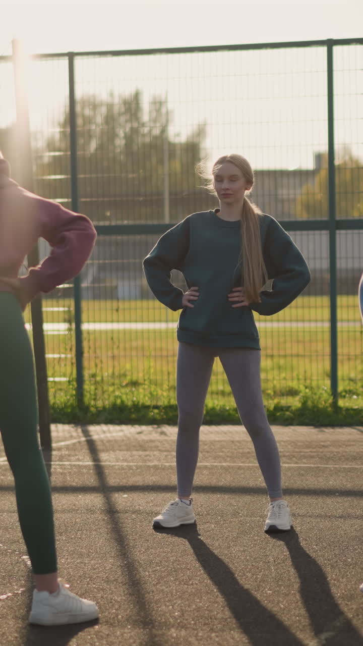 damas con leggings y capuchones participan en actividades de fitness en una cancha al aire libre con un edificio residencial y un campo de fútbol en el fondo, destacando el ejercicio físico y el trabajo en equipo