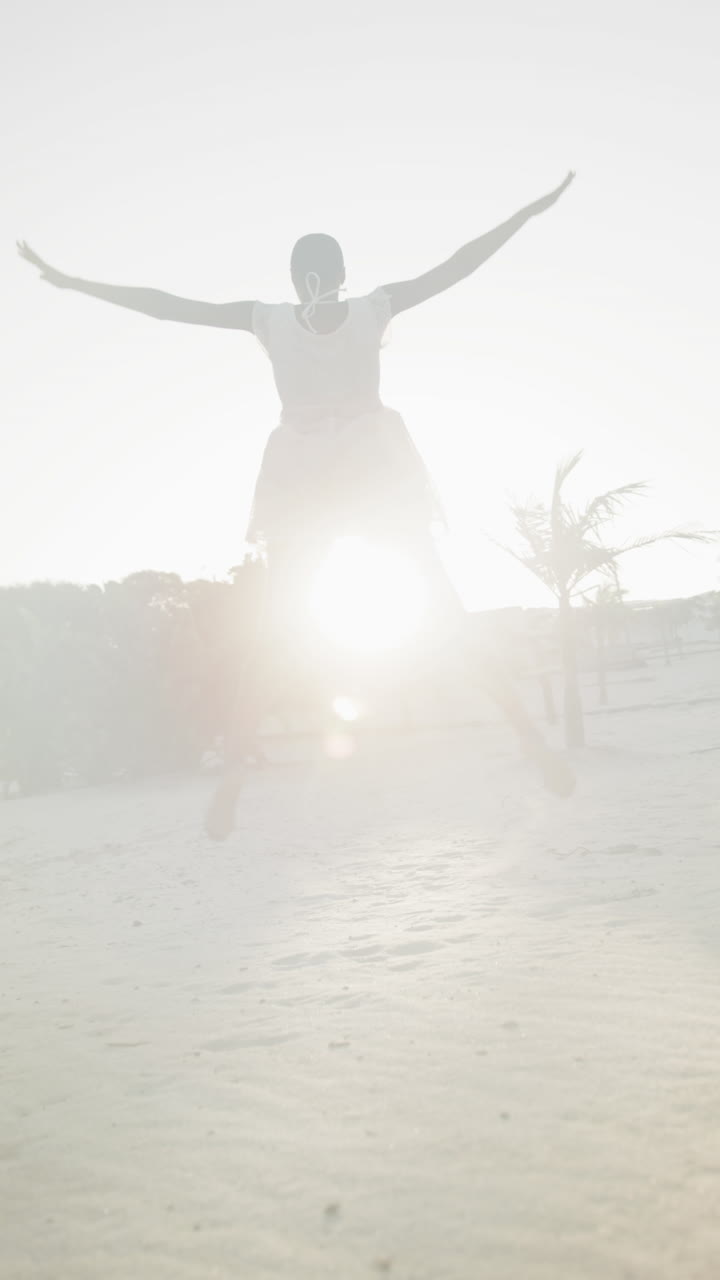 Vertical video of happy african american woman in dancing barefoot, backlit on beach, slow motion