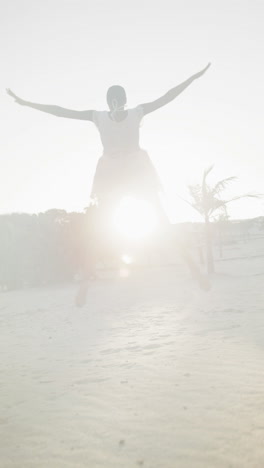 vertical video of happy african american woman in dancing barefoot, backlit on beach, slow motion