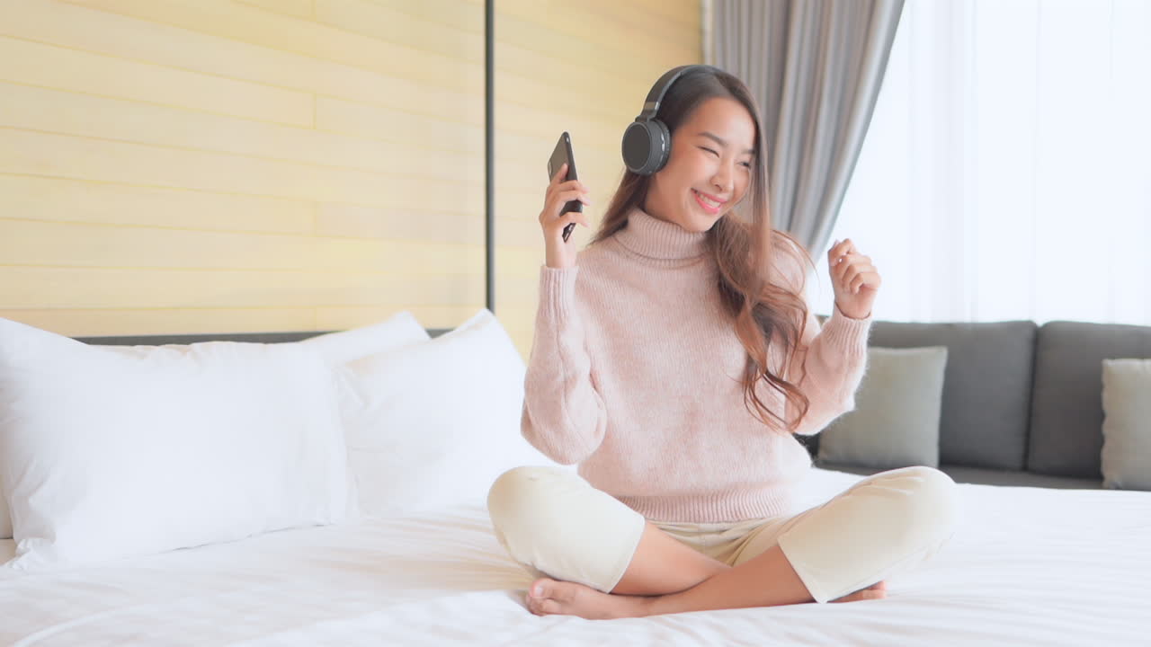 A lovely Asian girl wearing on-ear headphones is listening to music and dancing while holding her cellphone in hand, excited. Sitting on the bed with her legs crossed