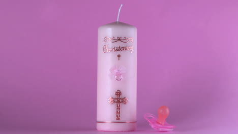 pink pacifier falling beside baptism candle on pink background