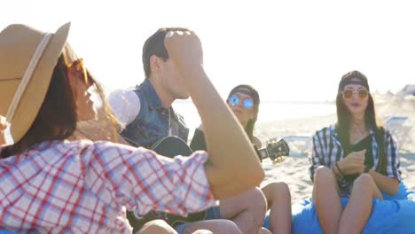 Young-man-playing-guitar-among-group-of-friends-sitting-on-easychairs-on-the-beach-and-singing-on-a-summer-evening.-Lens-flare.-Slowmotion-shot