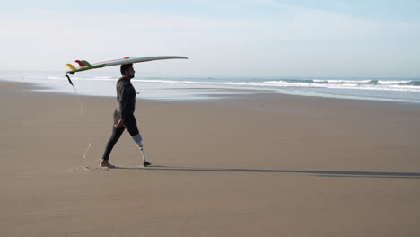 long shot of a male surfer with prosthetic leg walking on beach with surfboard on his head