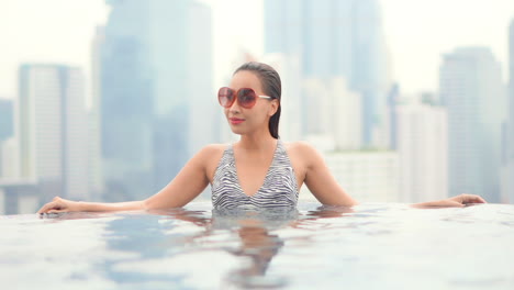 joven mujer asiática con gafas de sol en la piscina de la azotea urbana y rascacielos en segundo plano.