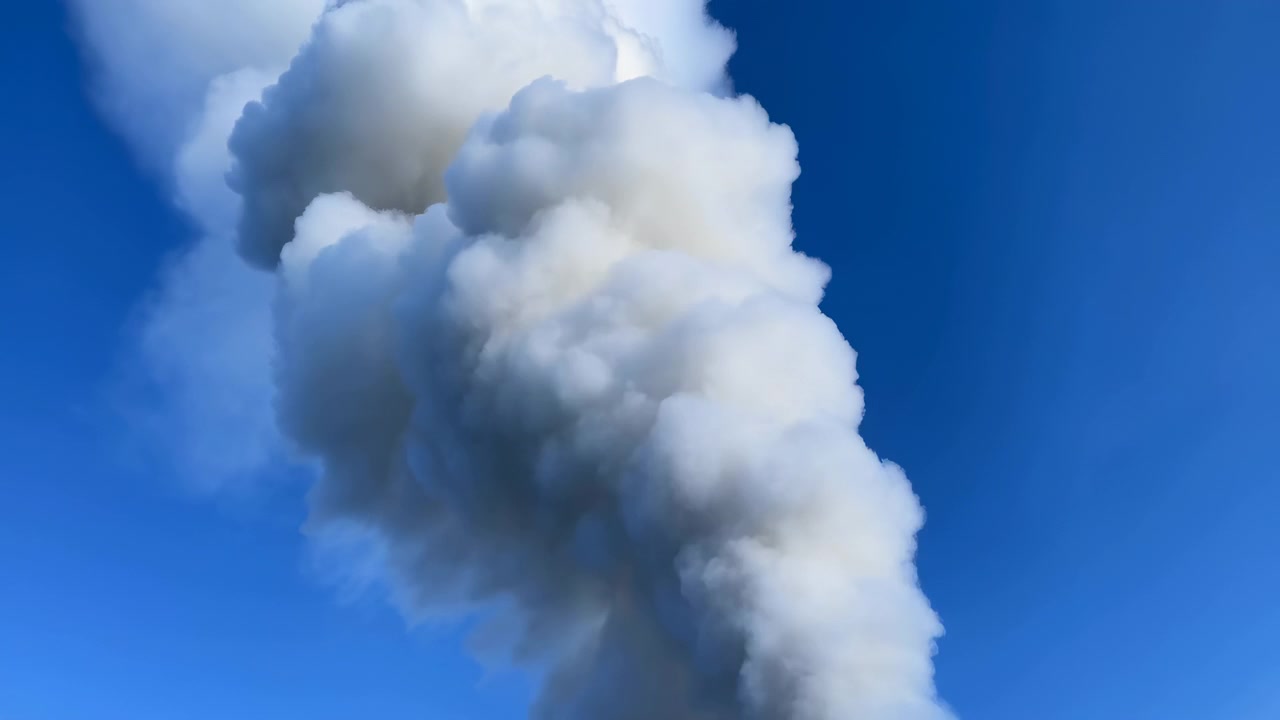En hög kolonn av tjockt vitt moln eller ånga stiger genom bildens mitt mot en livfullt blå himmel, med mjuka, skiktade böljor och starkt dagsljus.