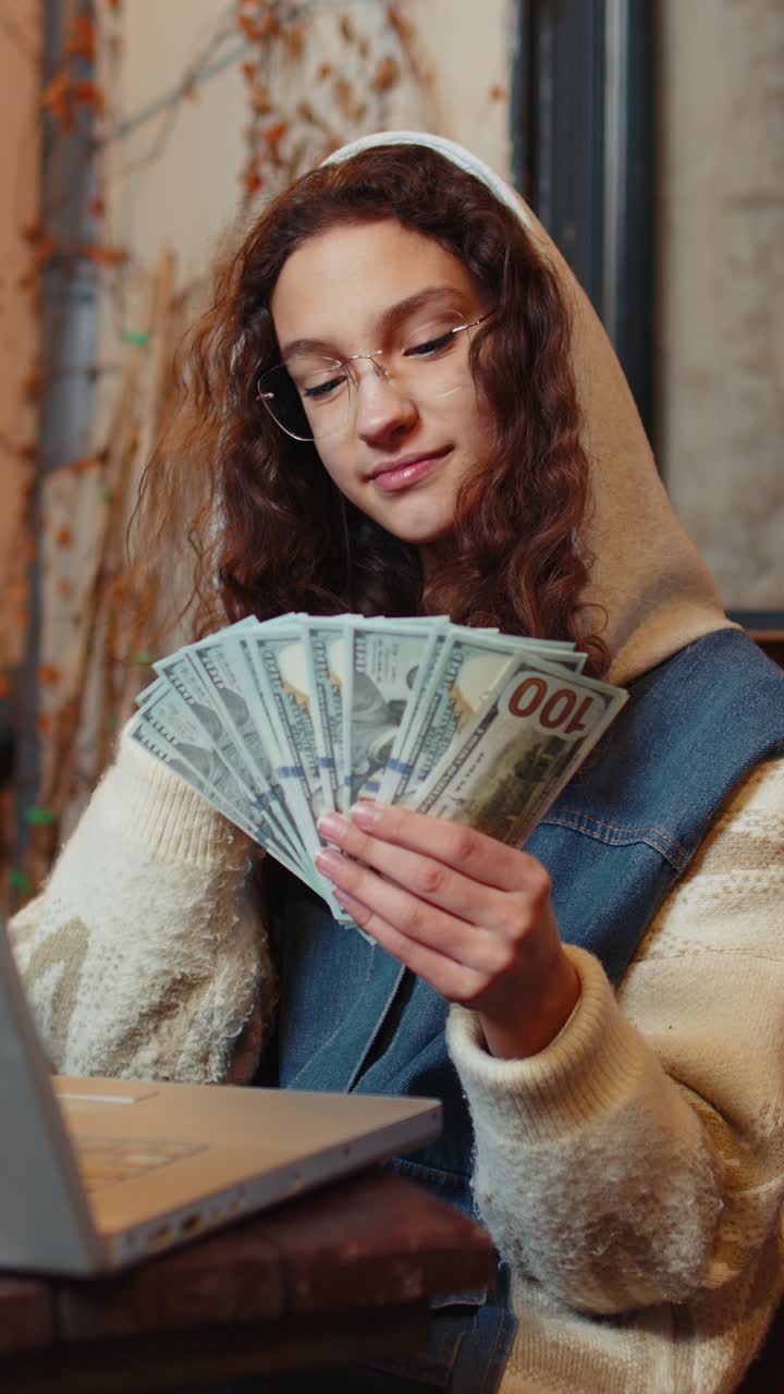 Happy rich girl using laptop winner victory holding fan of cash money in dollar banknotes in cafe