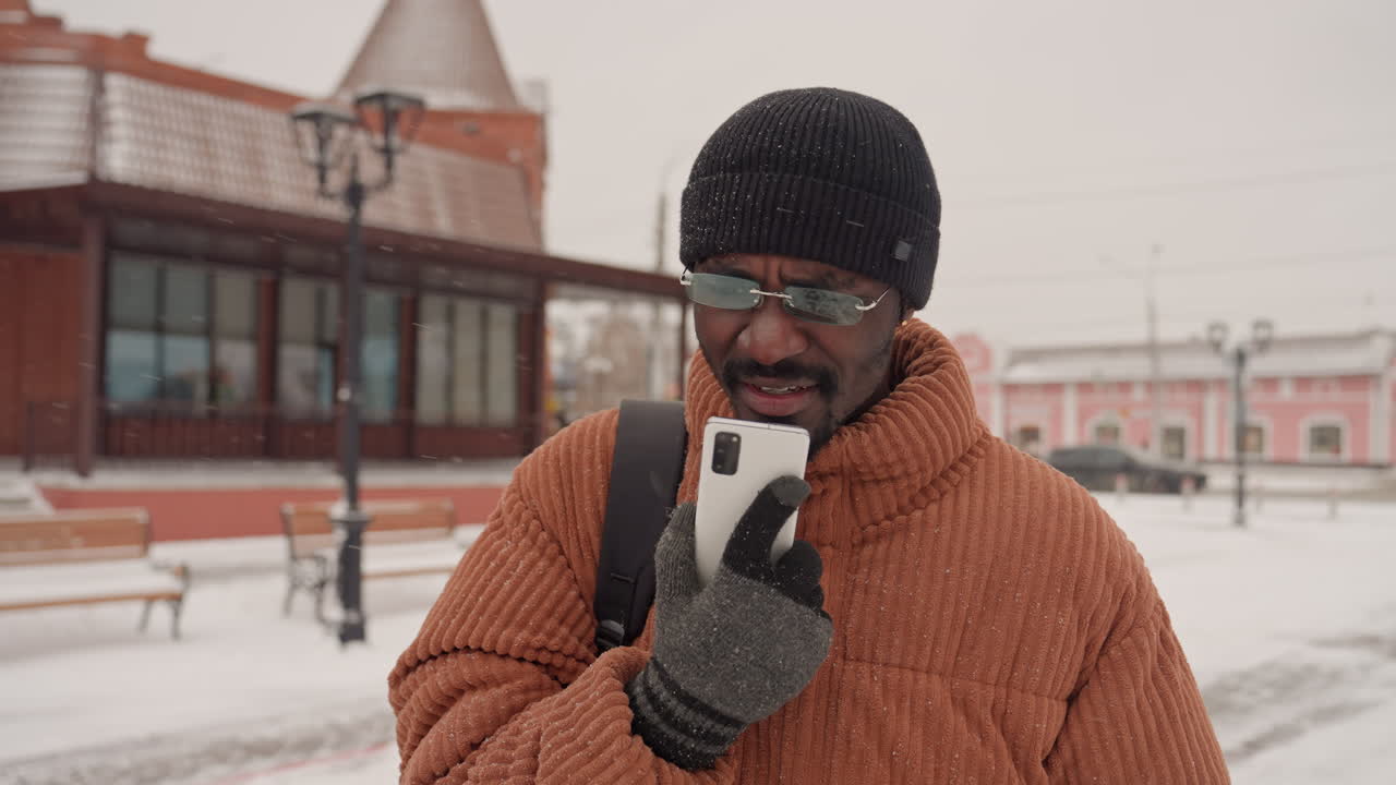 Uomo nero con cappotto arancione che parla al telefono in una piazza innevata, che indossa un berretto nero e guanti di lana, zaino in spalla, espressione pensierosa, nevicata leggera, panchine vuote e lampione, mattoni dai colori tenui