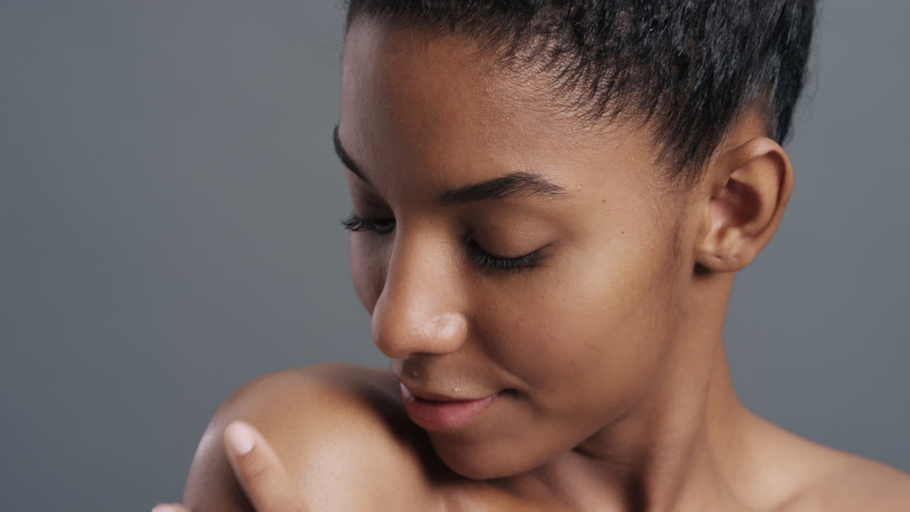retrato em close-up de uma bela mulher afro-americana tocando ombros nus com as mãos desfrutando de pele lisa e saudável perfeita beleza natural em fundo cinza