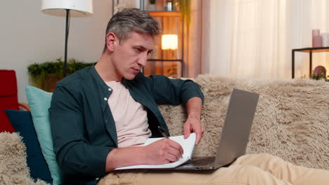 Young man takes notes in notebook while looking at laptop calm and focused on remote online work