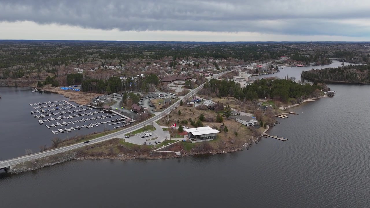 Kenora, Ontario. Drönarbild över Lake of the Woods som drar sig bort från den kanadensiska flaggan och avslöjar en marina, staden Kenora, en sjönära motorväg, bilar, båtar och träd en molnig vårdag.