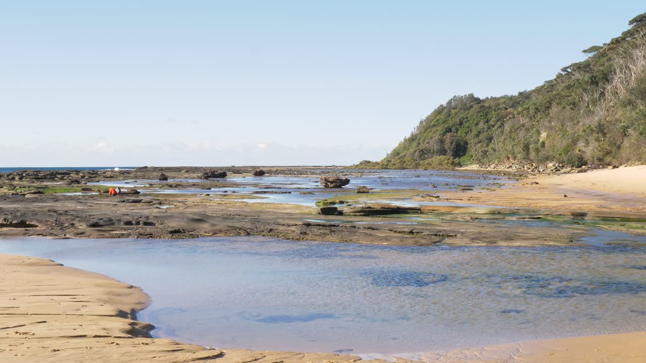 Slow motion scenic landscape showing ocean water flowing over rocky reef shoreline with forest bushland cliff headland and sweeping coastline views in the background Bateau Bay Central Coast Australia