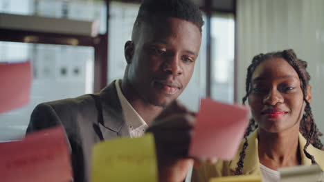 black man and woman write on post-it sticky note in office portrait through glass wall with sticks