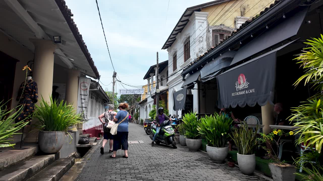 A bustling street scene in Galle Fort, Sri Lanka, featuring people strolling past quaint shops and motorcycles parked by the roadside. T
