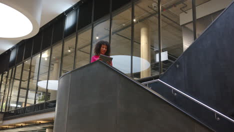 using tablet, businesswoman standing on staircase in modern office building