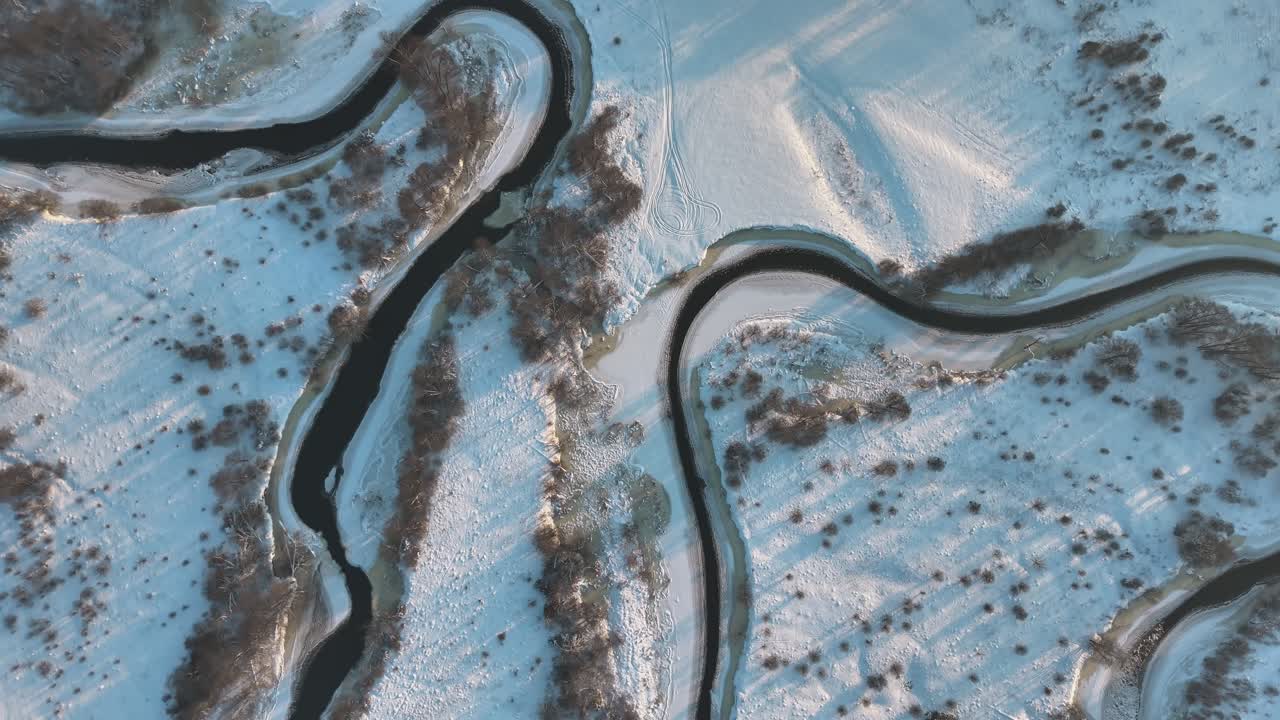 Top-down aerial view of a winding river cutting through snowy fields near Varena, Lithuania, highlighting abstract winter patterns and a peaceful frozen countryside landscape