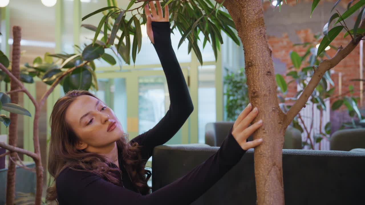 Young woman with long hair dressed in black reaches up to gently touch vibrant green leaves of indoor tree while surrounded by lush foliage and soft natural light in modern interior space