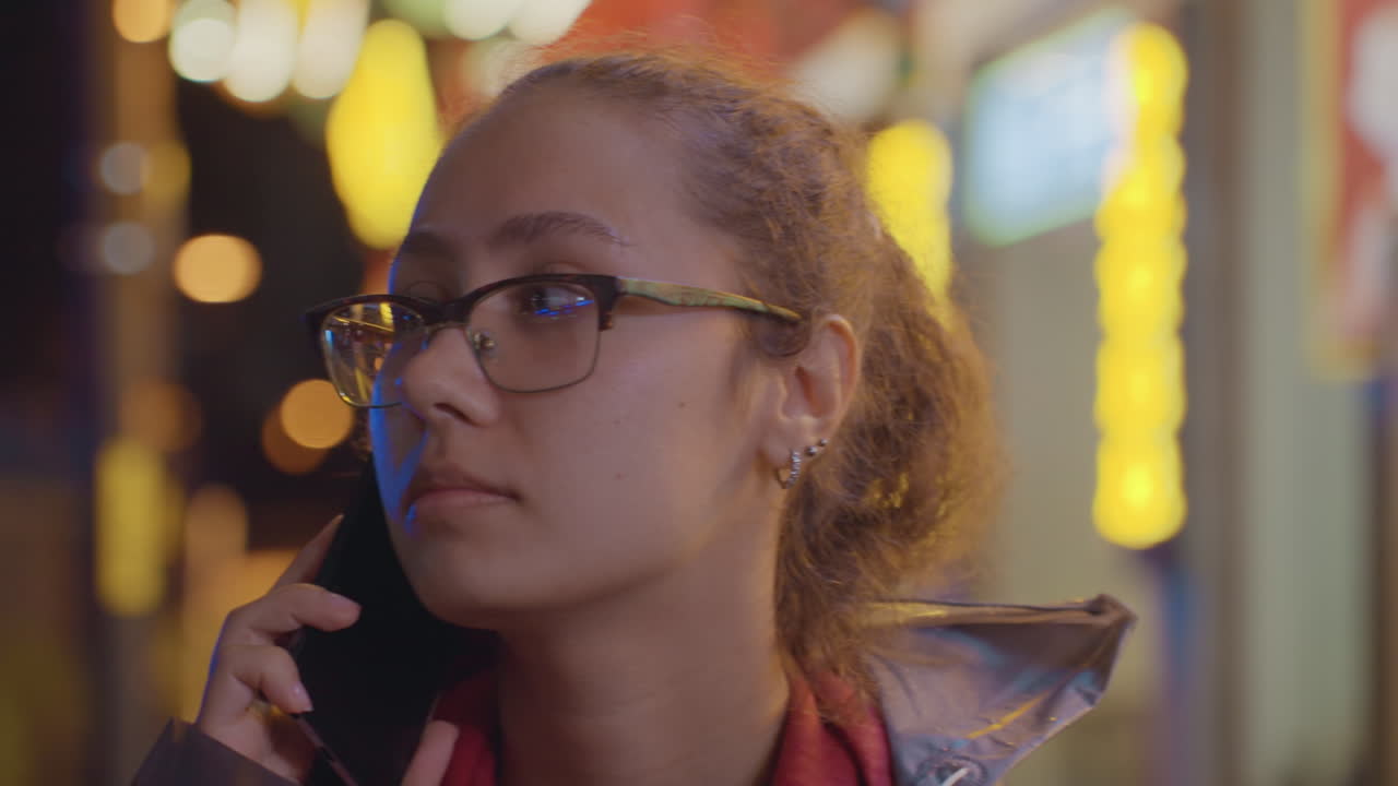 Jovem mulher ao telefone com bokeh de néon ao fundo, conversa noturna amigável com sorriso brincalhão e escuta atenta, óculos captando luzes quentes, jaqueta casual e cachecol, estilo de vida espontâneo.