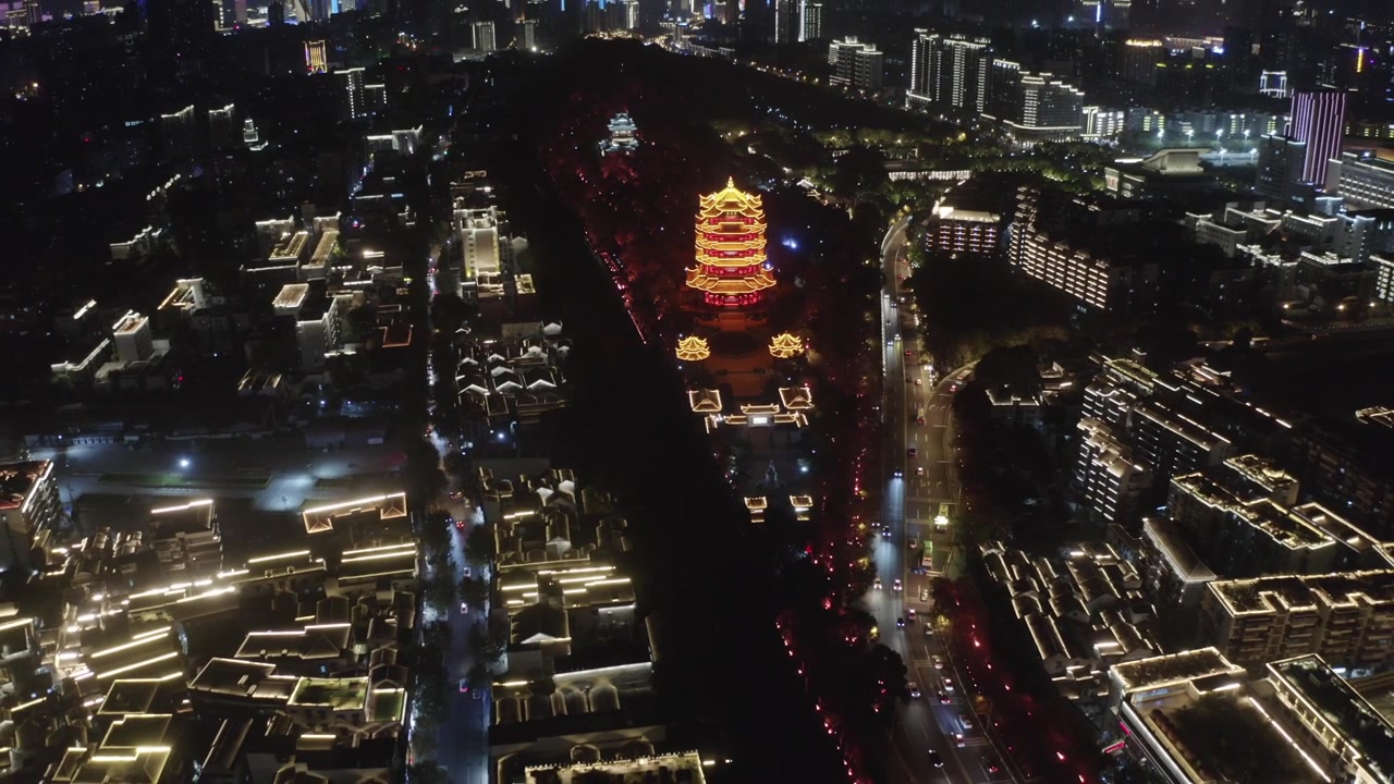Vista aérea de drone da icônica Torre do Grou Amarelo, intensamente iluminada e brilhando em vermelho dourado à noite, cercada por edifícios modernos da cidade e tráfego em Wuhan, província de Hubei, China.