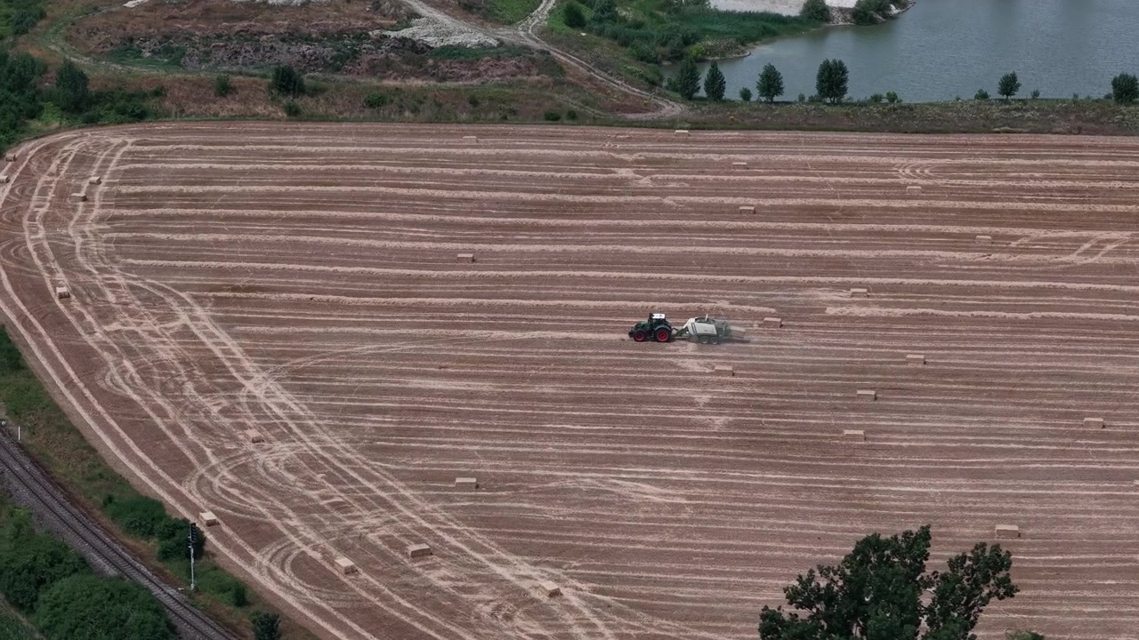 Una vista aerea mostra un trattore che imballa la paglia in un campo secco e raccolto con balle sparse, evidenziando le operazioni agricole estive