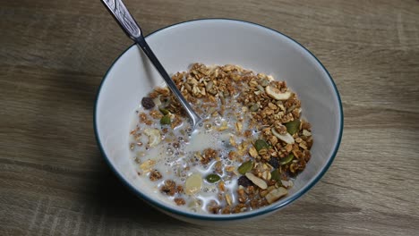 Granola-En-Un-Bol-Con-Una-Cuchara-Y-Leche-De-Almendras-Recién-Vertida-Haciendo-Burbujas,-Semillas,-Frutos-Secos,-Desayuno-Saludable