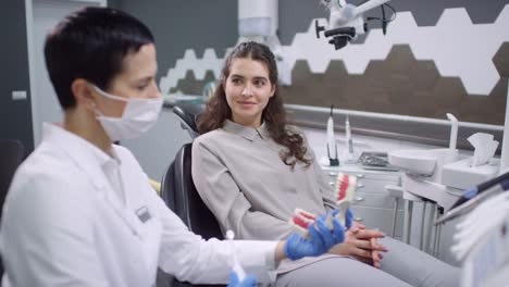 Mujer-Dentista-Con-Bata-Blanca-Y-Guantes-Usando-Cepillo-De-Dientes-Y-Mostrando-Al-Paciente-Cómo-Cepillarse-Los-Dientes-Correctamente-Usando-Dentaduras-Dentales